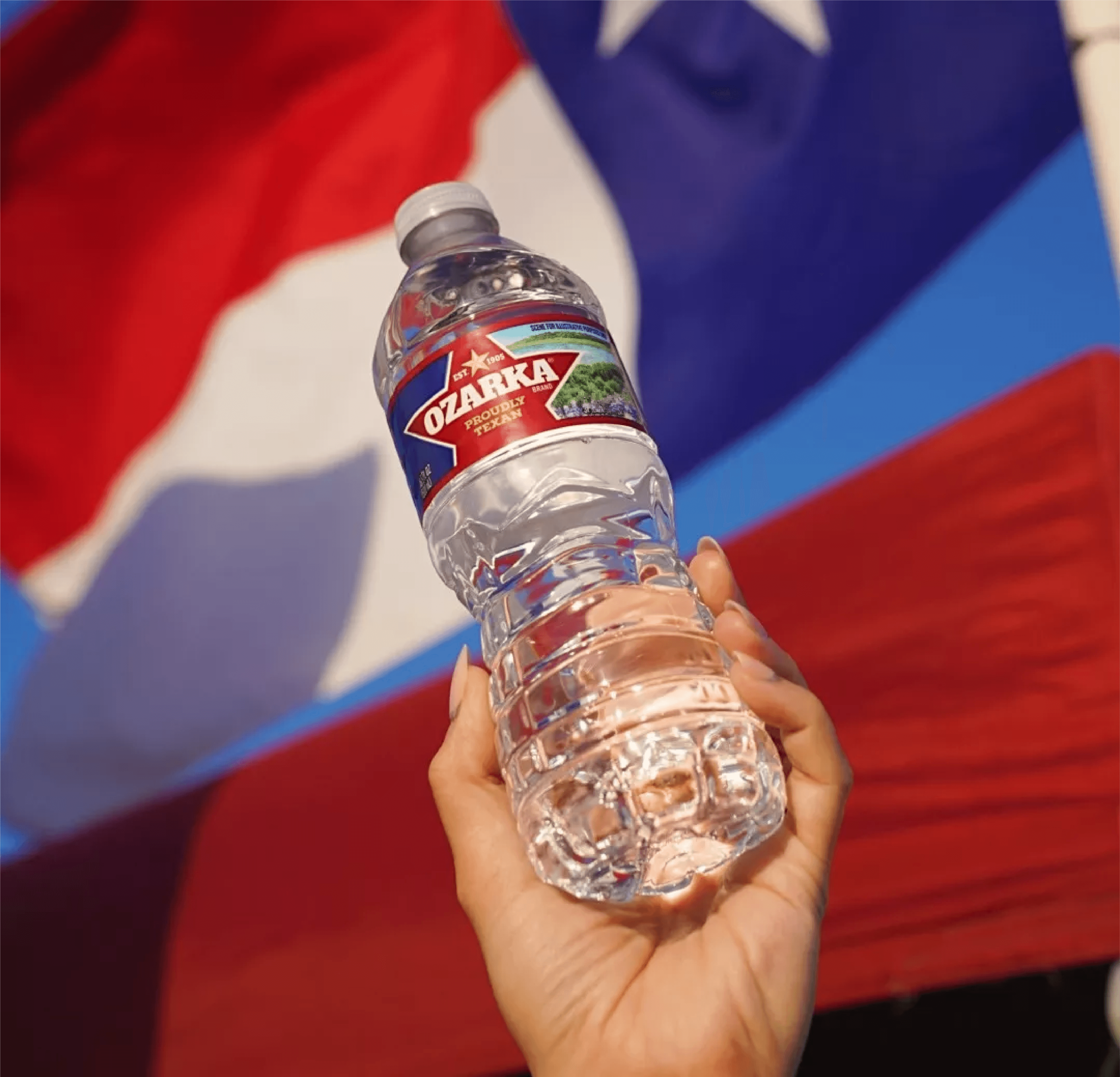 A hand holding an Ozarka water bottle in front of a waving Texas flag